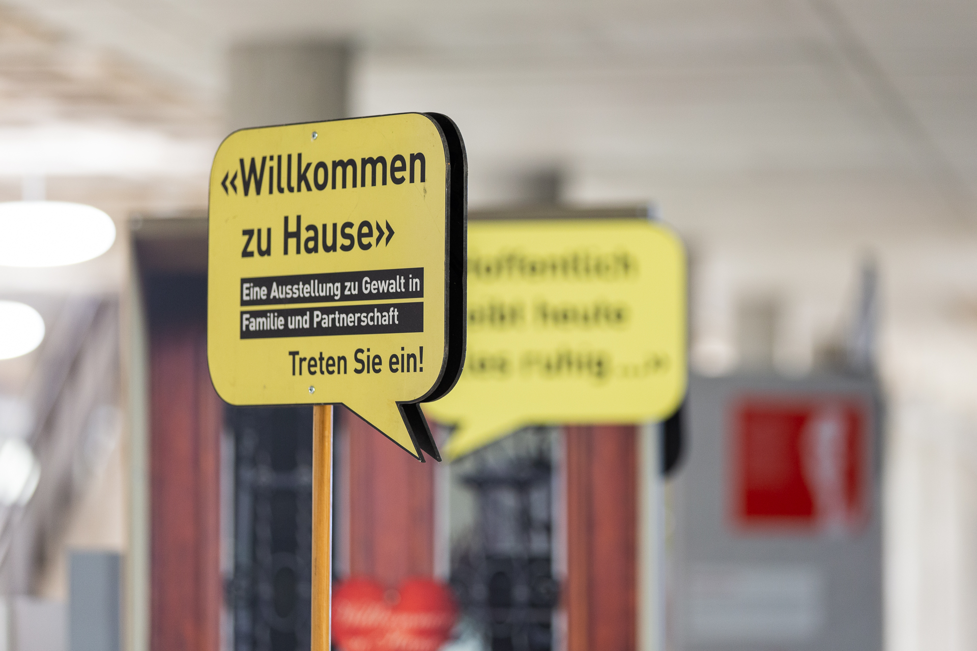 Ein gelbes Schild in Form einer Sprechblase steht in einem Innenraum. Darauf steht: «Willkommen zu Hause – Eine Ausstellung zu Gewalt in Familie und Partnerschaft. Treten Sie ein!». Im Hintergrund ist ein weiteres, unscharfes gelbes Schild zu sehen. Die Umgebung wirkt hell und modern, vermutlich Teil einer Ausstellung oder eines öffentlichen Gebäudes zum Thema häusliche Gewalt.